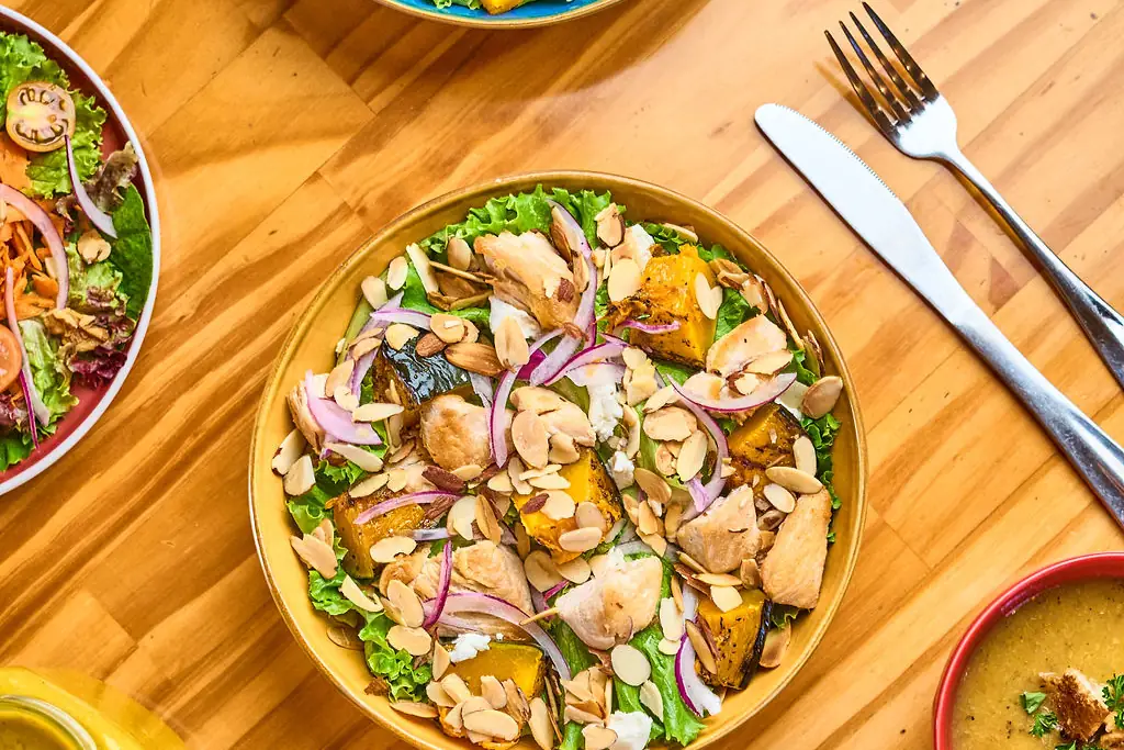 Ensalada con pollo, verdes y cebolla morada en un plato amarillo, vista desde arriba en una mesa de madera, junto a otra ensalada y una crema