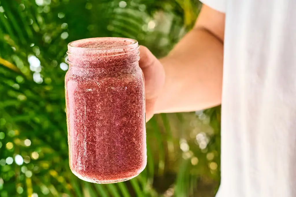 Batido morado servido en un frasco de vidrio, sostenido en la mano frente a plantas
