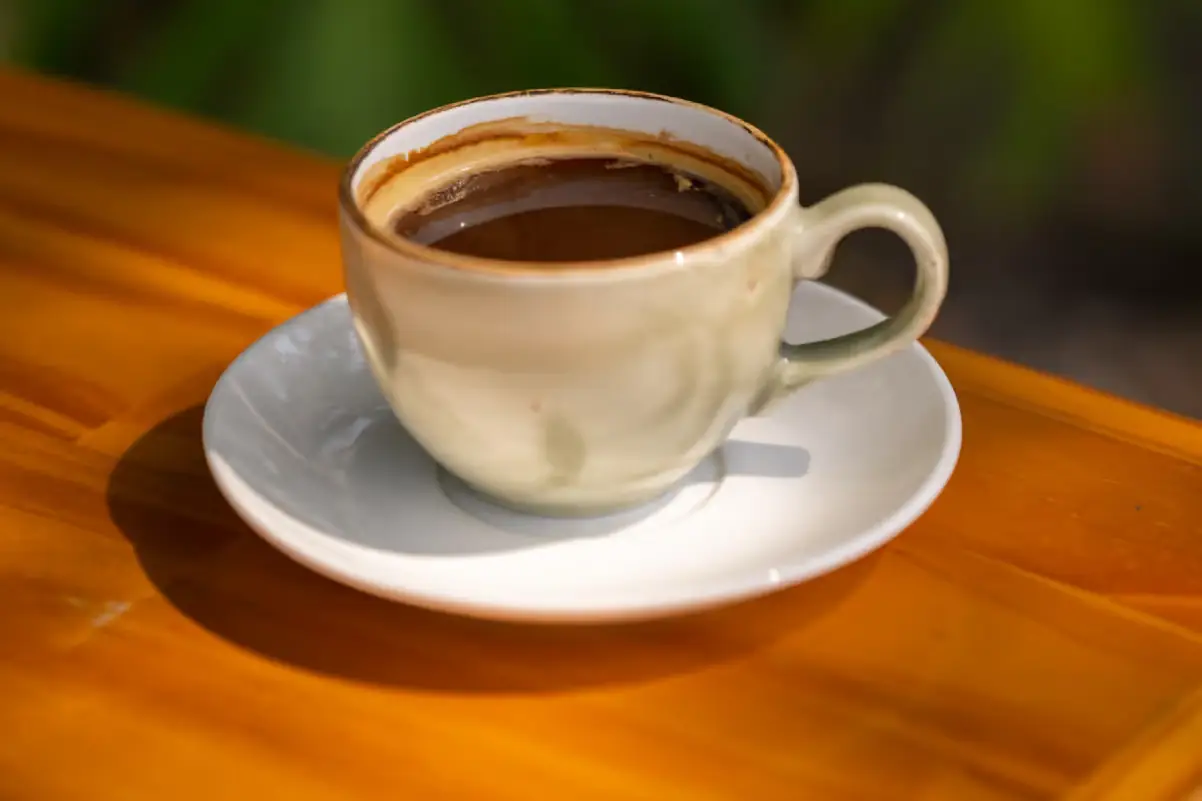 Taza de café negro en un plato blanco sobre una mesa de madera, con plantas al fondo