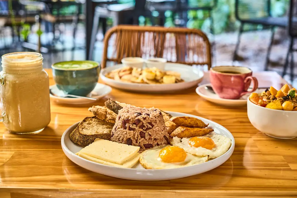 Bandeja Wö Kàpi con gallo pinto, huevos fritos, plátano maduro, queso fresco y pan, junto a un matcha, un batido y un café